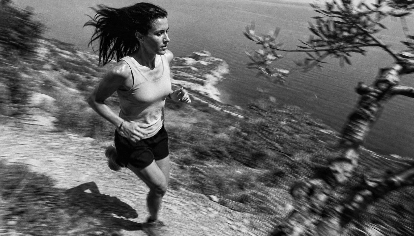 A woman running a rocky coastal trail past olive branches above the Mediterranean